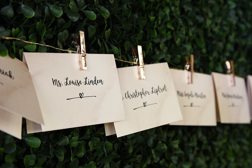 Place cards hanging on a gold wire string in front of a lush green wall | Vintage Garden Wedding at the Historic Alfred I DuPont Building in Miami, FL